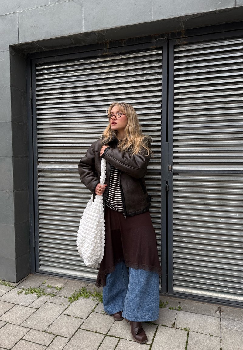 Jeune femme portant des lunettes, une veste sombre, un haut rayé, une jupe superposée et un jean large, tenant un grand sac blanc texturé devant une porte métallique à volet.