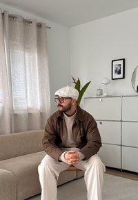 Un homme portant une casquette blanche, une veste marron et des lunettes est assis, les mains jointes, dans un salon beige près d'une plante et d'un meuble blanc.