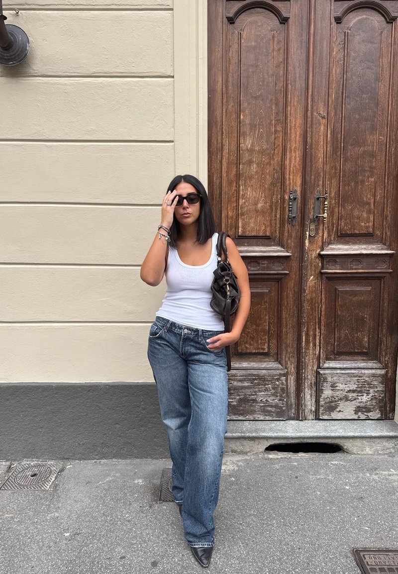 Camisola branca sem mangas, jeans de perna larga azul-claro, bolsa preta e óculos de sol; de pé contra portas de madeira com painéis elaborados.