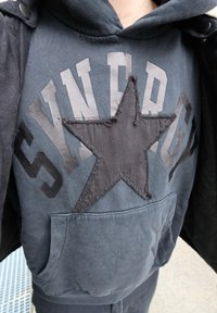 Gros plan d'une personne portant un sweat à capuche sombre avec le mot "STANFORD" et une grande étoile noire cousue sur la zone de la poche avant.