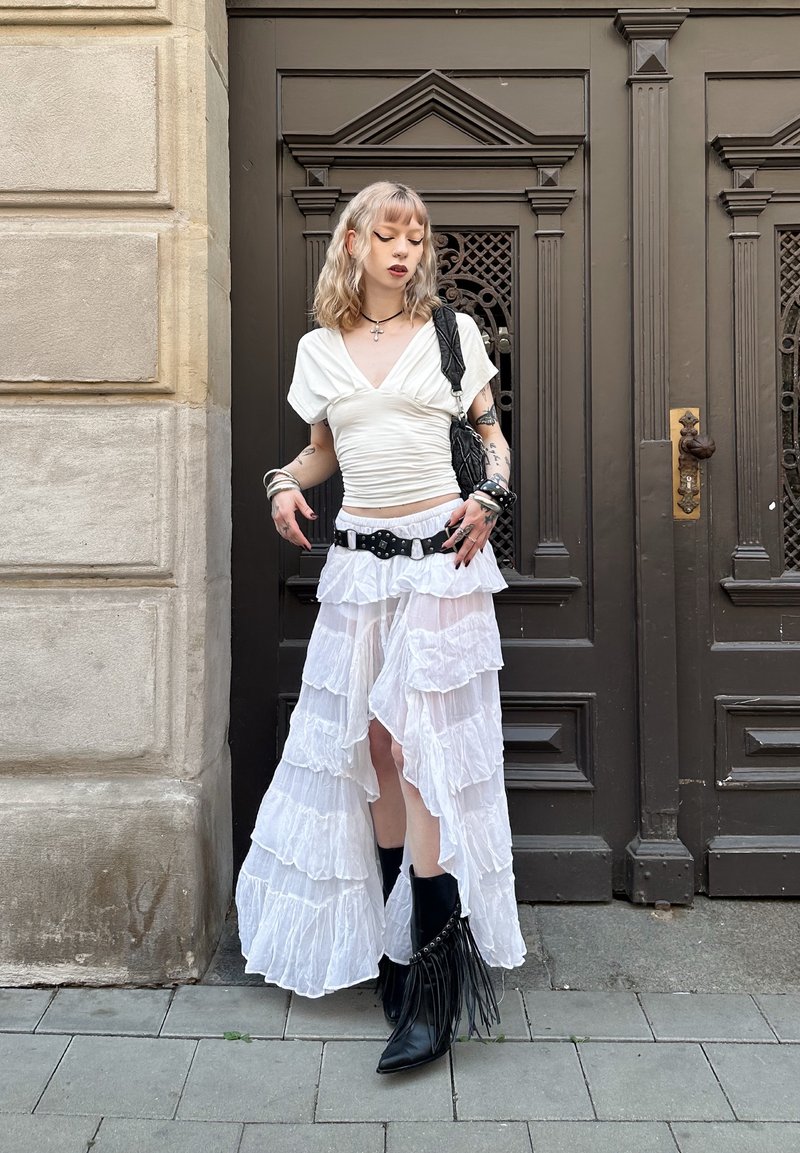 Jeune personne aux cheveux clairs et tatouages portant un haut blanc froncé, une jupe blanche superposée, une ceinture noire et des bottes noires à franges, debout près d'une porte sombre.