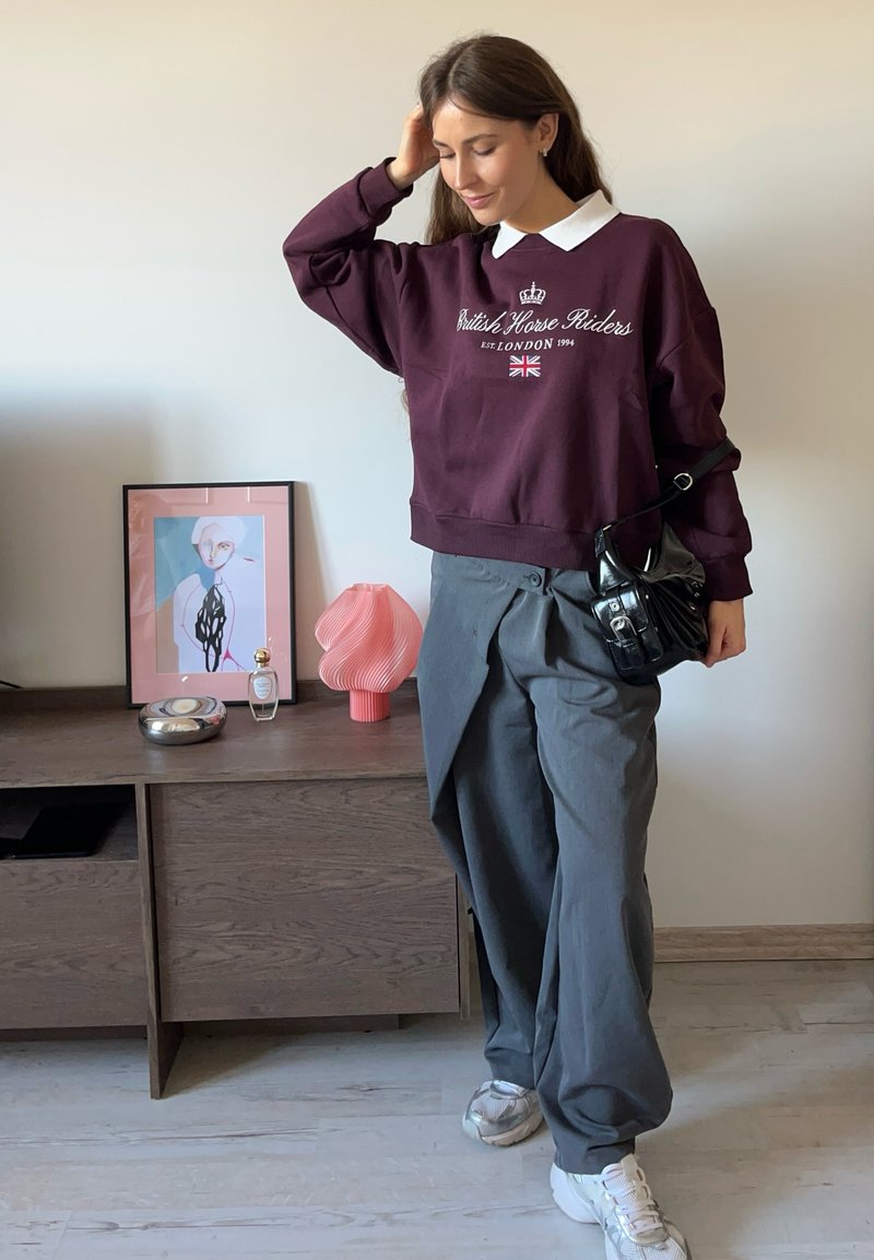 Felpa marrone con collo bianco, pantaloni grigi a gamba larga e scarpe da ginnastica bianche. Borsa nera con dettagli. Texture morbide e stile casual.