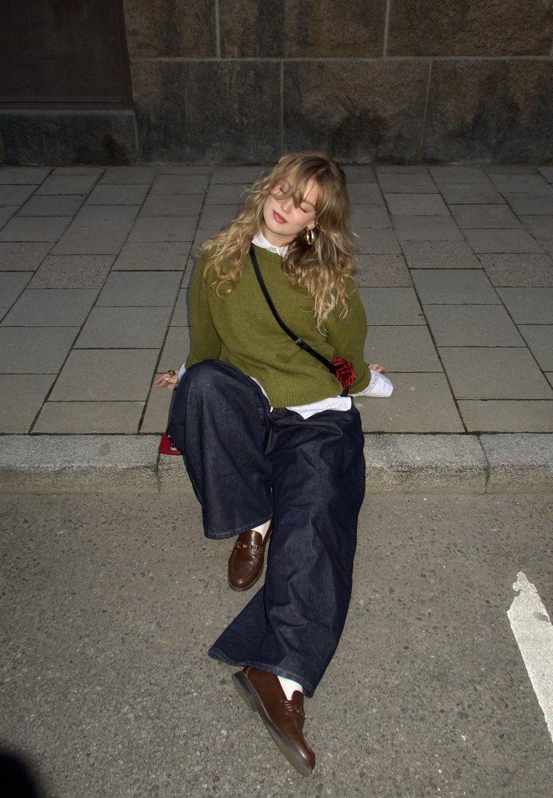 Jeune femme aux cheveux bouclés blonds assise sur le trottoir, portant un pull vert, un pantalon large foncé, des chaussures marron et un sac bandoulière la nuit.