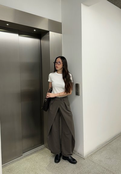 Mujer con cabello largo y oscuro y gafas, de pie junto a puertas de ascensor cerradas, vistiendo camisa blanca, pantalones anchos color oliva, zapatos negros y sosteniendo un bolso negro.