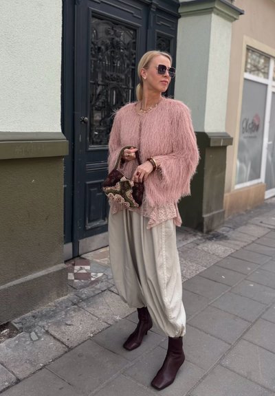 Mujer con chaqueta peluda rosa, pantalones beige holgados, botas marrones y gafas de sol, sosteniendo un bolso bordado mientras está de pie en la acera de una ciudad.