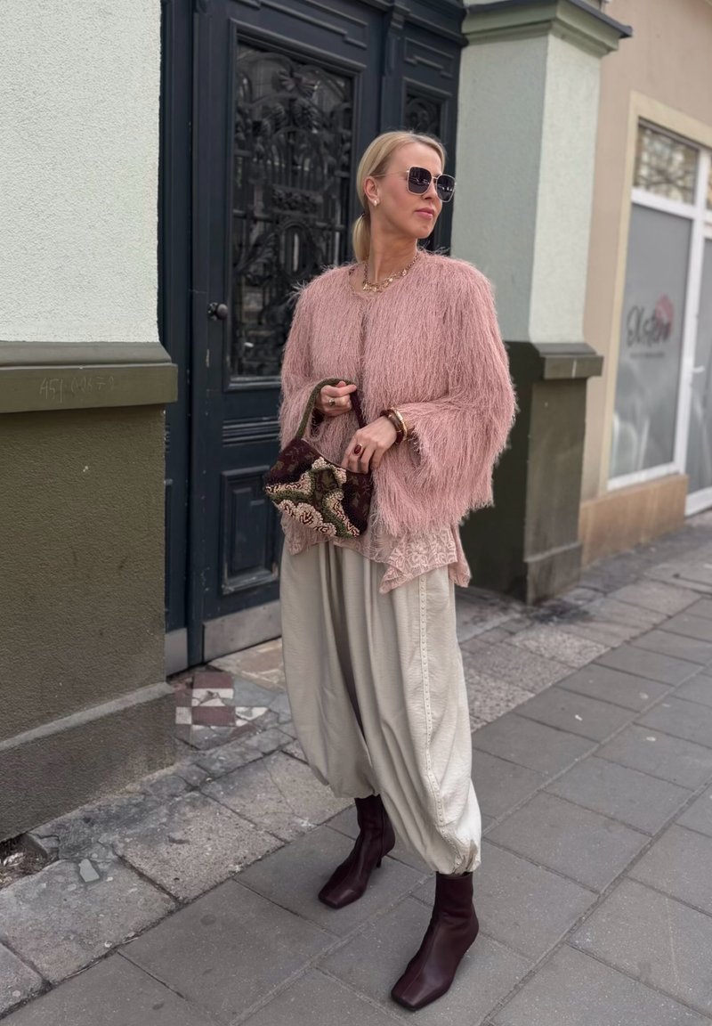 Frau in einer rosa flauschigen Jacke, lockeren beigen Hosen, braunen Stiefeln und Sonnenbrille, die eine bestickte Tasche hält und auf einem Bürgersteig in der Stadt steht.