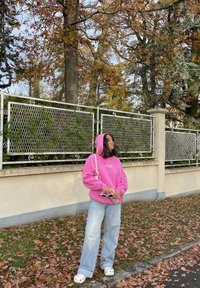 Per didelė rožinė megztinė, šviesiai mėlyni plačiakojai džinsai, balti saulės akiniai, balta rankinė ir balti crocsai. Fone matyti metalinė tvora ir rudeniniai lapai.