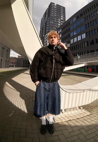 Chaqueta negra corta con cremallera y bolsillos, combinada con shorts de mezclilla hasta la rodilla. Medias blancas y zapatos negros completan el atuendo. Entorno urbano.