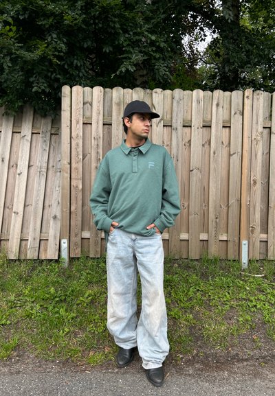 Camisa polo verde de manga larga con cuello y logo, combinada con unos vaqueros holgados de lavado claro y botas negras, con un fondo de cerca de madera.