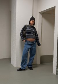 Pull gris rayé recadré, jean bleu ample, chaussures noires et sac à main marron. Porte un bonnet, des écouteurs sont visibles.