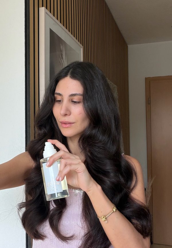 Frau mit langen dunklen Haaren hält eine klare Flasche mit weißer Pumpe und sprüht daraus, steht drinnen nahe einer Wand mit Holzvertäfelung und eingerahmter Dekoration.