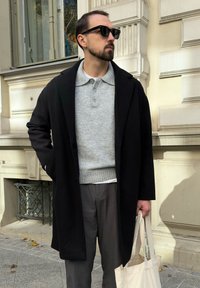 Vīrietis melnā uzvalkā, pelēkā džemperī un saulesbrillēs stāv ārā, turot rokās beidzamā krāsā ir tirgus soma pie krējuma ēkas fasādes.