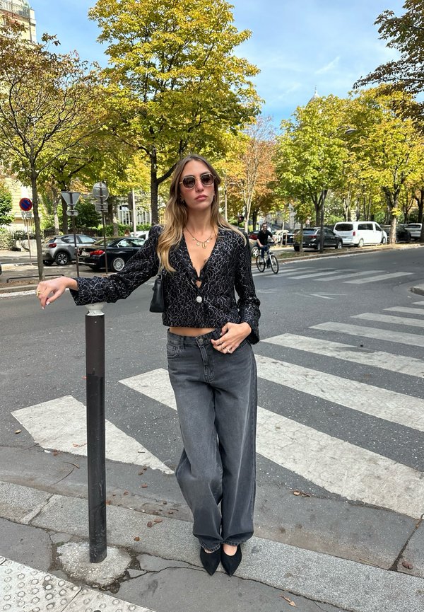 Mujer con gafas de sol y blusa negra con estampado se apoya en un poste cerca de un paso de peatones en una calle de la ciudad con coches y árboles al fondo.