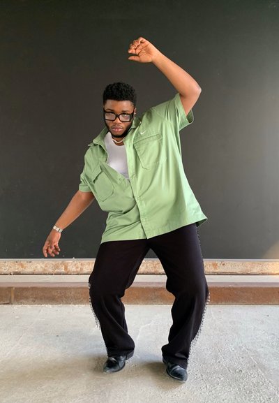 Camisa verde de manga corta con botones, camiseta blanca debajo, pantalones negros con flecos, zapatos negros. Persona adopta una pose dinámica contra un fondo oscuro.
