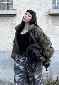 Veste en fausse fourrure couleur sable, haut noir, pantalon camouflage et sac noir texturé. Les détails remarquables comprennent une bordure en dentelle et des accents de bijoux en argent.