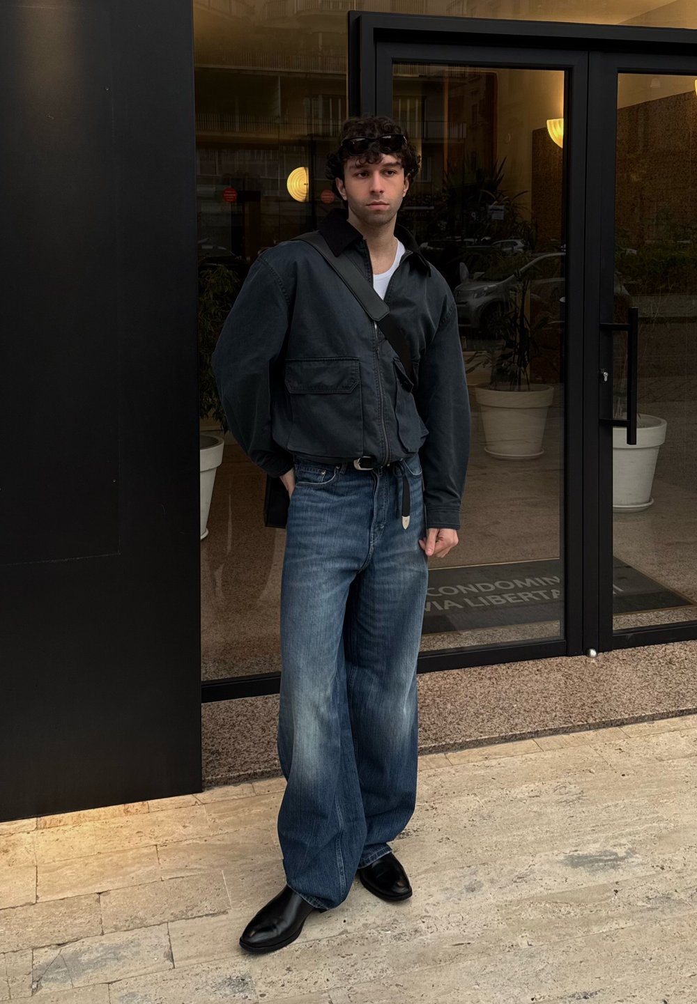 Jeune homme aux cheveux bouclés portant une veste noire, un large jean bleu, des chaussures noires et un sac bandoulière noir, debout devant des portes vitrées.