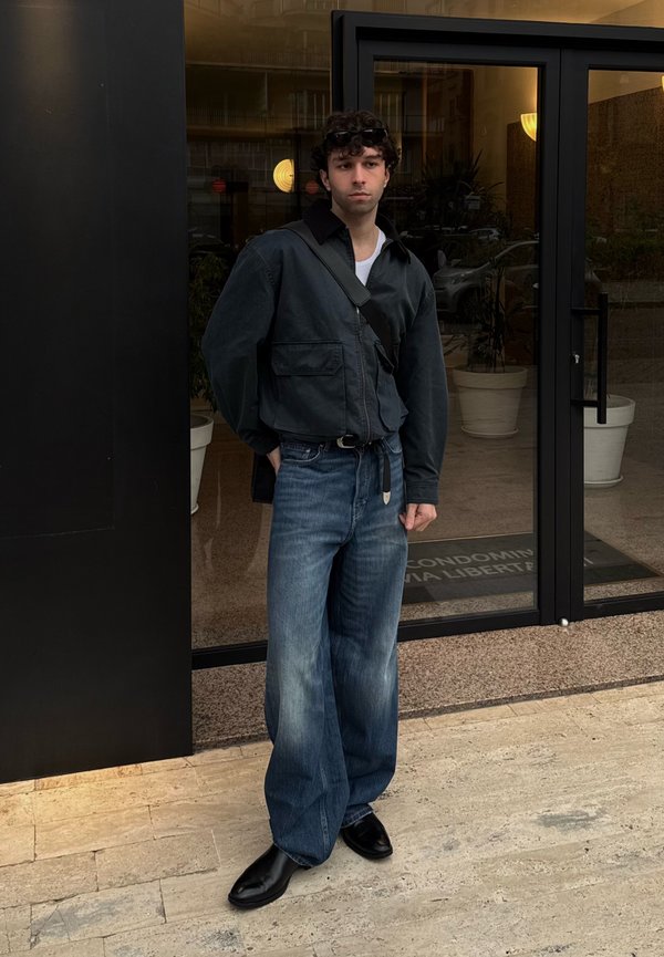 Giovane uomo con capelli ricci che indossa una giacca nera, jeans blu larghi, scarpe nere e porta una borsa a tracolla nera, in piedi davanti a porte di vetro.