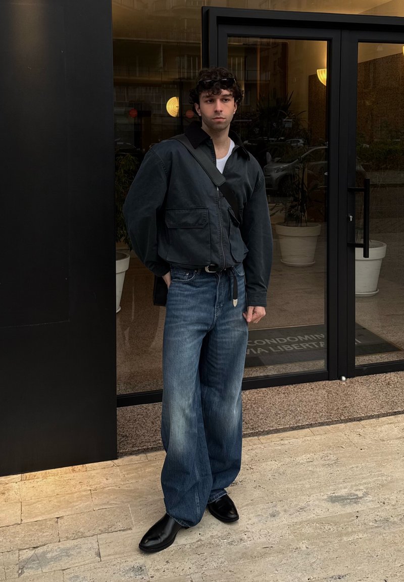 Giovane uomo con capelli ricci che indossa una giacca nera, jeans larghi blu, scarpe nere e porta una borsa a tracolla nera, in piedi davanti a porte di vetro.