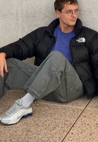 Giubbotto a sacco nero, maglietta blu a collo rotondo, pantaloni cargo verdi e scarpe da ginnastica grigie con accenti blu; seduto su una superficie di pietra testurizzata.