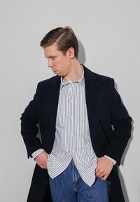 Άνδρας που φοράει μαύρο παλτό πάνω από ριγωτό πουκάμισο με κουμπιά και μπλε τζιν, κοιτάζοντας κάτω με τα χέρια στις τσέπες, μπροστά σε ένα απλό γκρι φόντο.