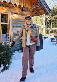 Donkergroen opgeblazen jack, grijze trui, bruine cargobroek, zwarte handschoenen, zonnebril en een rode schoudertas. Besneeuwde buitenomgeving met een houten hut.