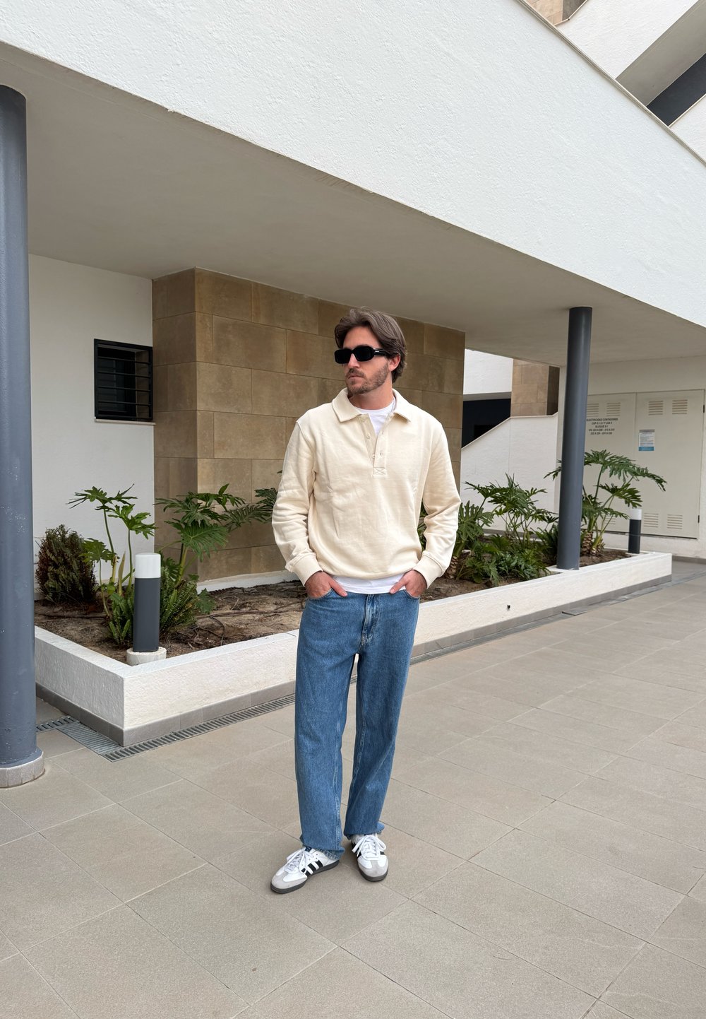 Conjunto casual com um pulôver creme, t-shirt branca, jeans azuis e sapatilhas brancas. O fundo inclui plantas e arquitetura moderna.
