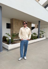 Conjunto casual com um pulôver creme, t-shirt branca, jeans azuis e sapatilhas brancas. O fundo inclui plantas e arquitetura moderna.