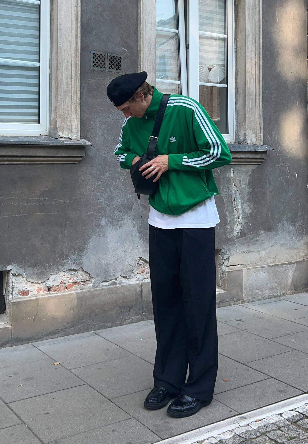 Zielona kurtka dresowa Adidas z białymi paskami, czarna torba, luźne czarne spodnie i czarne buty. Mężczyzna przegląda torbę, stojąc na chodniku.
