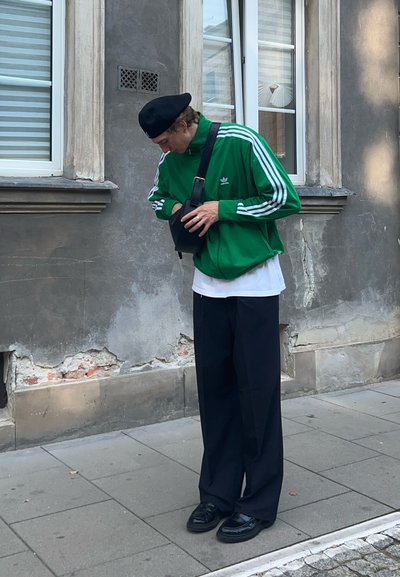 Chaqueta de chándal verde de Adidas con rayas blancas, bolsa negra, pantalones negros holgados y zapatos negros. Hombre mirando dentro de la bolsa, de pie en una acera.