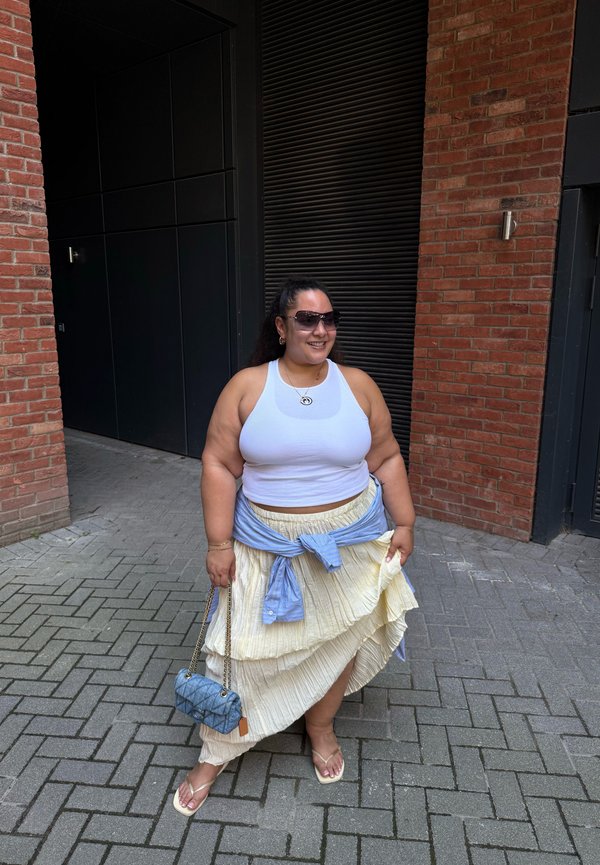 Femme portant un débardeur blanc, une jupe plissée crème, des lunettes de soleil, une chemise bleue nouée autour de la taille, tenant un sac à main bleu, debout dans une rue pavée.