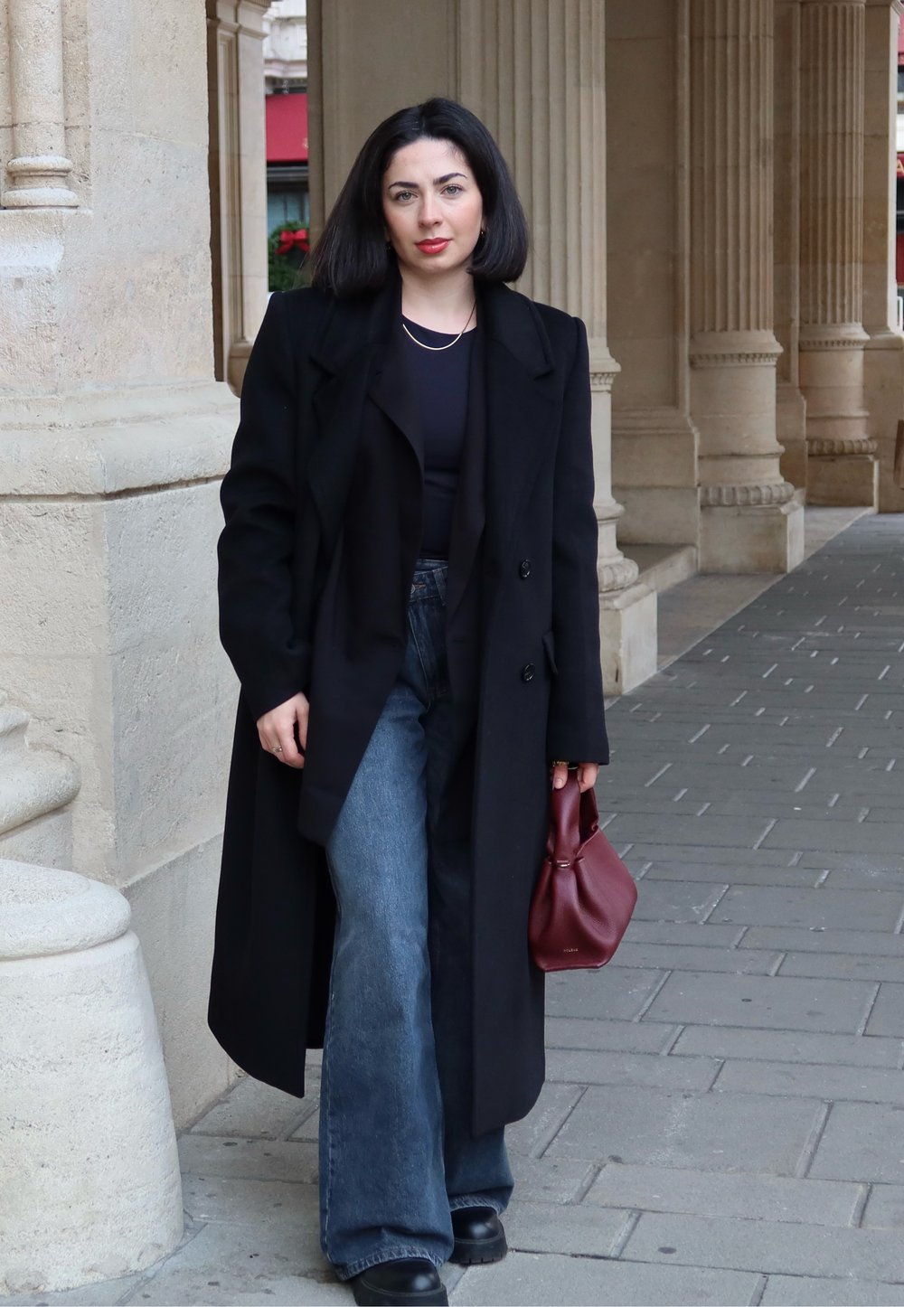 Zwarte overjas, zwart topje, blauw uitlopende jeans met hoge taille, zwarte plateau laarzen, een wijnrode handtas vasthoudend, staand op een stenen trottoir.