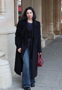 Zwarte overjas, zwart topje, blauw uitlopende jeans met hoge taille, zwarte plateau laarzen, een wijnrode handtas vasthoudend, staand op een stenen trottoir.