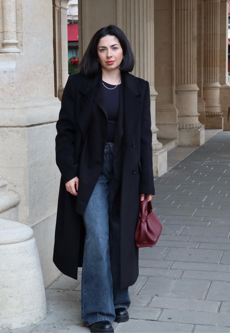 Sobretudo preto, camisa preta, jeans azuis de cintura alta e boca larga, botas pretas de plataforma, a segurar uma mala bordeaux, em cima de pavimento de pedra.