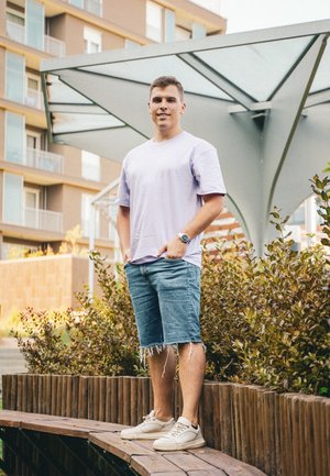Camiseta corta de manga corta en púrpura claro, pantalones cortos de mezclilla con bordes deshilachados, zapatillas blancas; de pie al aire libre cerca de follaje verde y una estructura de madera.