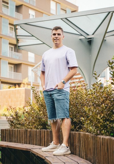 Camiseta corta de manga corta en púrpura claro, pantalones cortos de mezclilla con bordes deshilachados, zapatillas blancas; de pie al aire libre cerca de follaje verde y una estructura de madera.