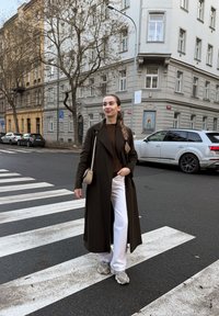 Long manteau marron, pull marron foncé, pantalon blanc et baskets grises. Passage pour piétons au premier plan, bâtiments et voitures garées en arrière-plan.