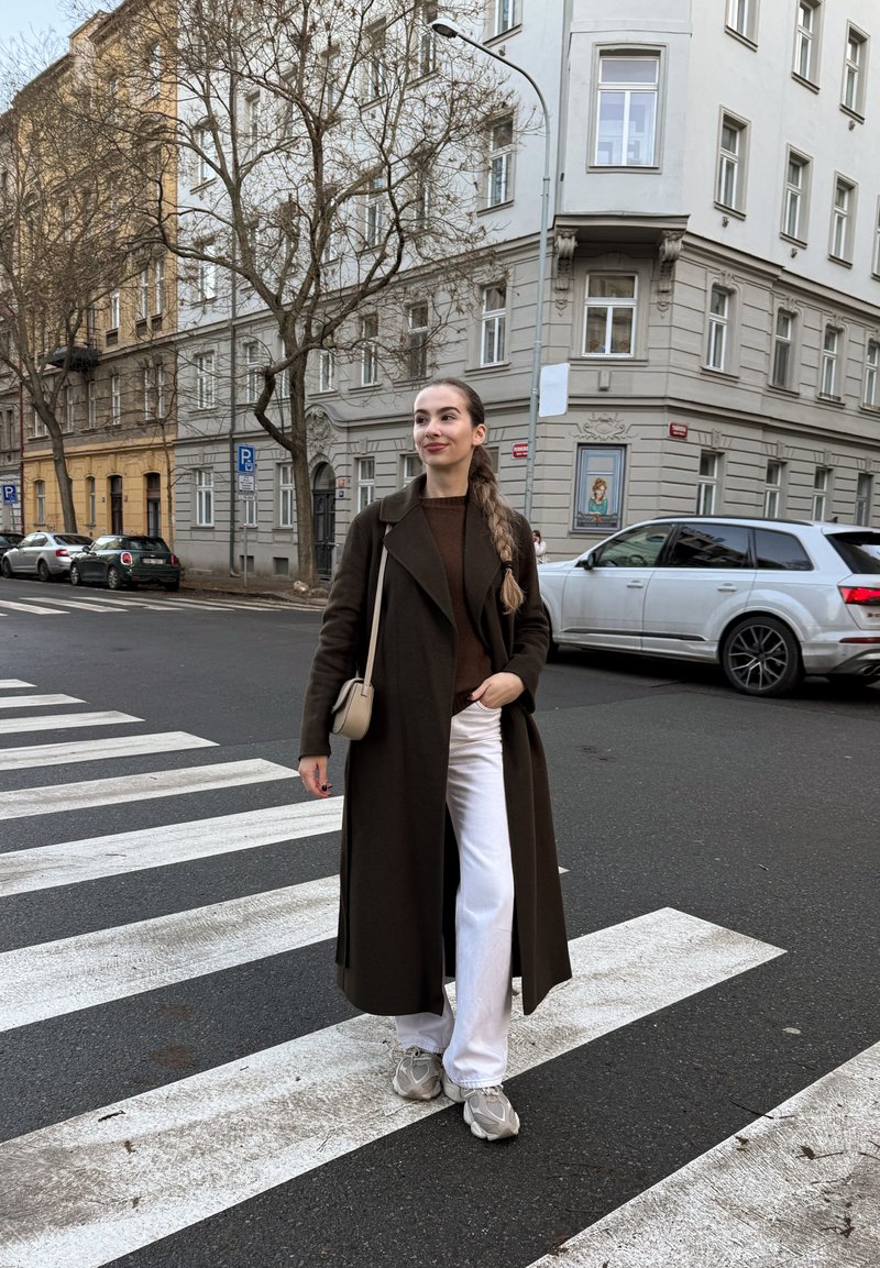 Lungo cappotto marrone, maglione marrone scuro, pantaloni bianchi e scarpe da ginnastica grigie. Strisce pedonali in primo piano, edifici e auto parcheggiate sullo sfondo.