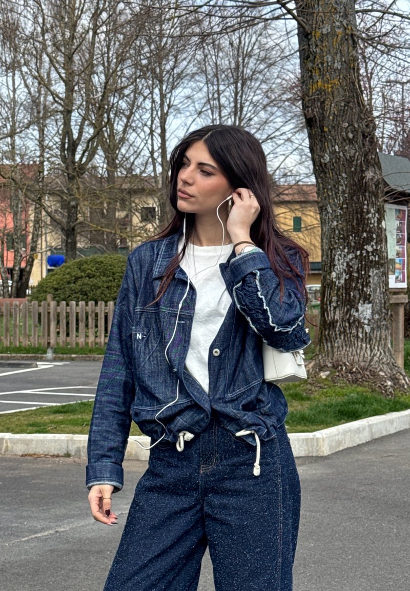 Jeune femme portant une veste et un pantalon en jean ajustant des écouteurs blancs en plein air, près des arbres et des bâtiments par une journée nuageuse.