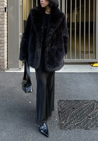 Personne portant un manteau en fausse fourrure noire, une longue jupe noire, des chaussures noires à bouts pointus et tenant un sac à main noir dans une rue urbaine.