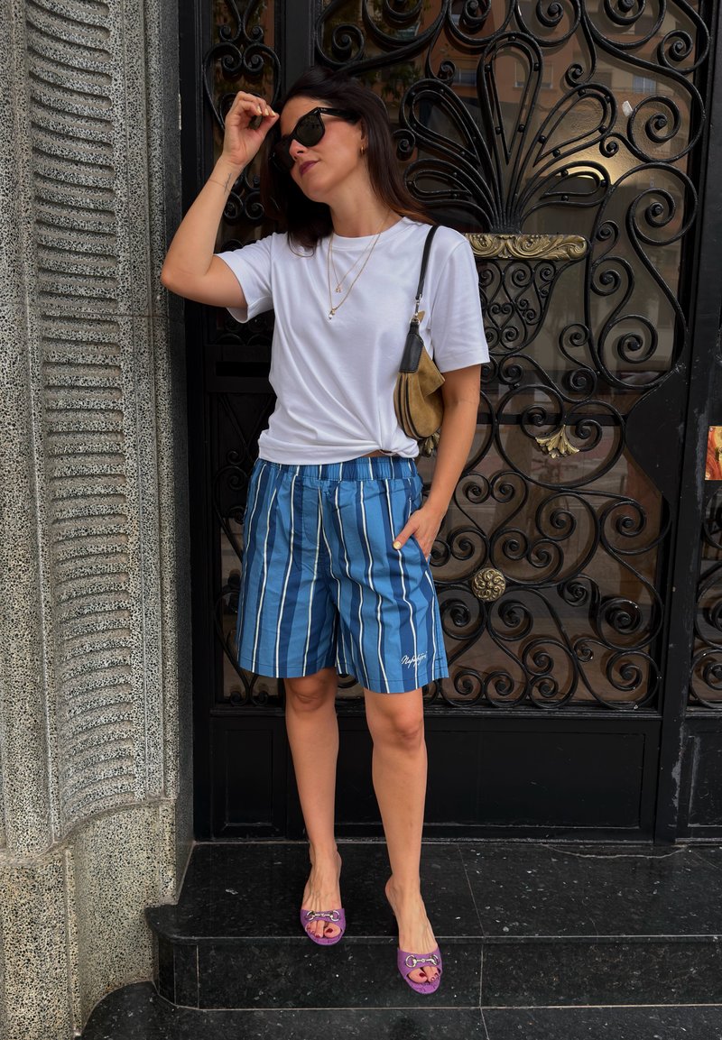 Femme ajustant ses lunettes de soleil, portant un t-shirt blanc, un short bleu rayé, des chaussures violettes et portant un sac à épaule marron, debout près d'une porte noire ornée.