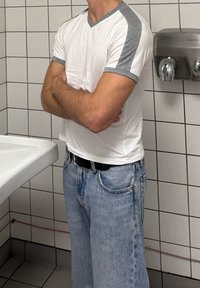 Άνδρας με λευκό και γκρι κοντομάνικο πουκάμισο και μπλε τζιν στέκεται με σταυρωμένα τα χέρια σε πλακακιασμένο μπάνιο κοντά σε νιπτή ρα και στεγνωτήρα χεριών.