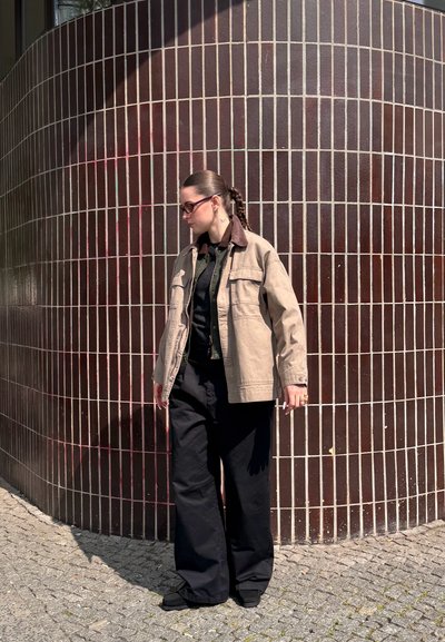 Chaqueta de lona beige con un cuello marrón, camiseta negra y pantalones anchos negros, de pie contra una pared de azulejos oscuros. Lleva puestas unas gafas.