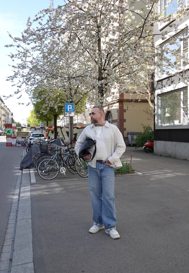 Homme portant des lunettes de soleil, une chemise blanche et un jean, debout sur le trottoir tenant une veste sous un cerisier en fleurs près de vélos stationnés.
