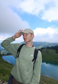 Mladý muž v zelené mikině Nike a béžové čepici jde na túru s batohem poblíž horského jezera pod polojasnou oblohou.