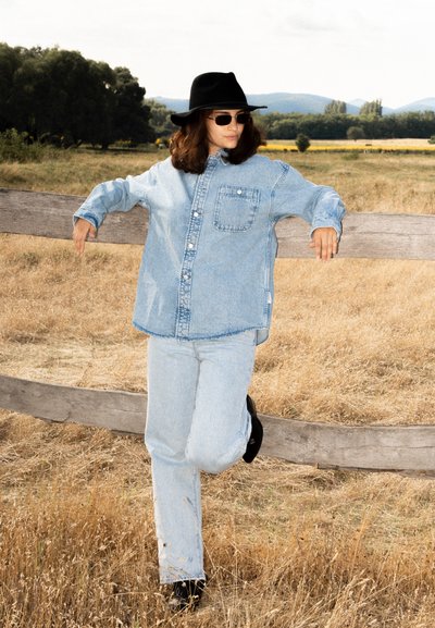 Camisa de denim azul claro y jeans a juego, con botones en la parte delantera y un bolsillo en el pecho. Un sombrero negro de ala ancha y gafas de sol completan el conjunto.