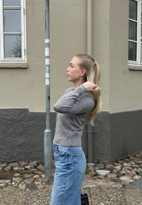 Cardigan grigio a maglia sopra una gonna di jeans blu, con lunghi capelli biondi raccolti. In piedi accanto a un muro grigio e a un palo della strada.