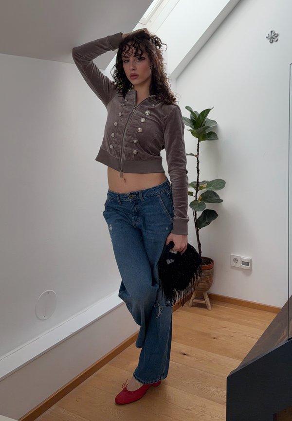 Woman with curly hair poses indoors wearing a cropped brown jacket, blue jeans, red shoes, holding a black fringed bag, standing near a potted plant.