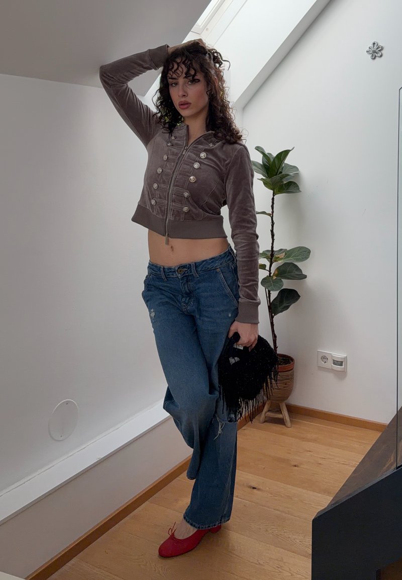 Frau mit lockigem Haar posiert drinnen, trägt eine kurze braune Jacke, blaue Jeans, rote Schuhe, hält eine schwarze Tasche mit Fransen und steht neben einer Topfpflanze.