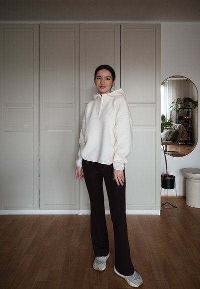 Mujer de pie en el interior vistiendo una sudadera blanca con capucha, pantalones negros y zapatillas blancas, con un espejo y plantas en macetas en el fondo.