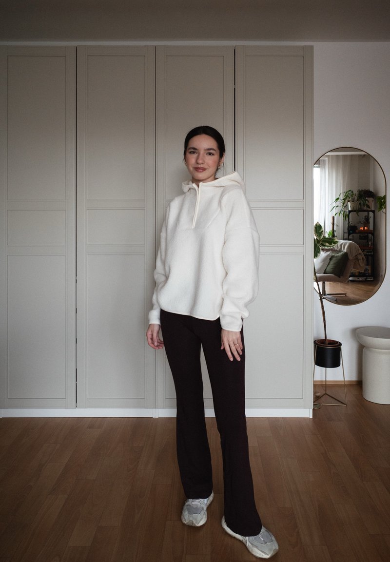 Γυναίκα στέκεται μέσα σε εσωτερικό χώρο φορώντας λευκή μπλούζα με κουκούλα, μαύρο παντελόνι και λευκά αθλητικά παπούτσια, με έναν καθρέφτη και φυτά σε γλάστρες στο παρασκήνιο.